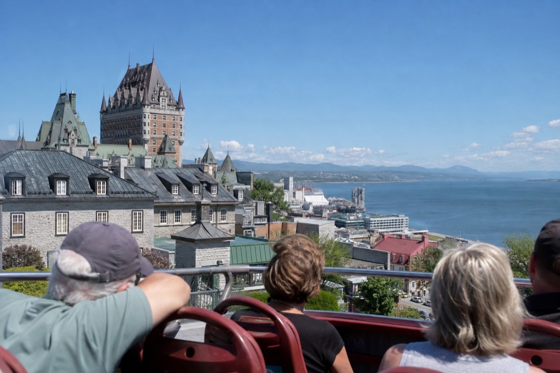 Passengers on the open upper deck of a sightseeing bus looking out over rooftops and the St. Lawrence River