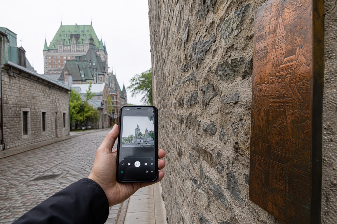Walker holding a phone with an audio tour beside a stone wall in Old Quebec, choosing between free and guided walking tours